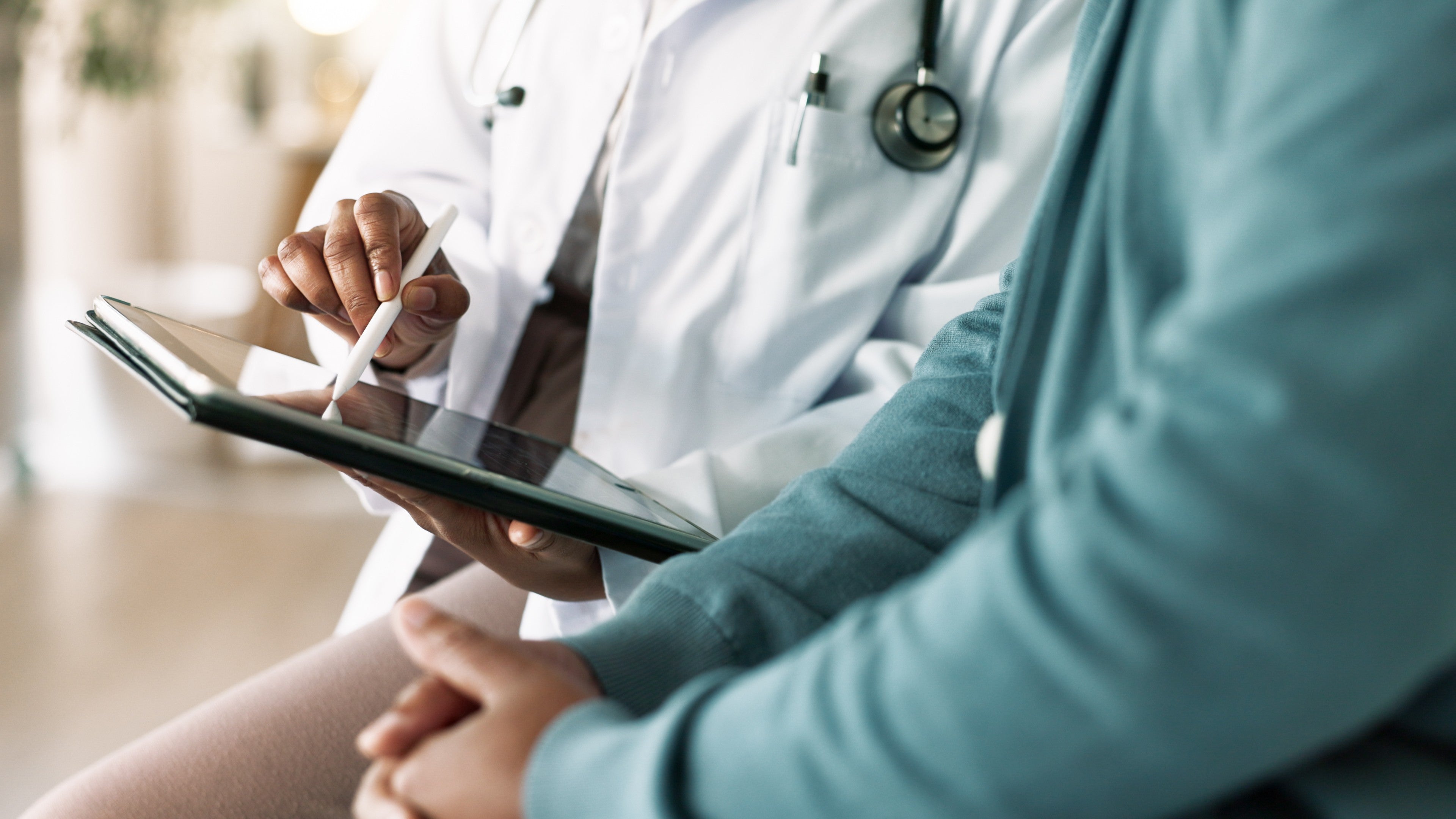 This image shows a close-up scene of a medical consultation between a healthcare professional and a patient.

On the left side, a doctor is partially visible, wearing a white coat with a stethoscope draped around their neck. Their face is not shown, but their hands are clearly visible. The doctor is holding a tablet (a flat touchscreen device) in one hand and using a white stylus pen to point at or write something on the screen.

On the right side, the patient is sitting close by, wearing a teal or light blue long-sleeve top. Only their torso and hands are visible. Their hands are gently clasped together in their lap, suggesting they are listening attentively.

The doctor appears to be explaining or reviewing information on the tablet, possibly test results, a treatment plan, or health data, while the patient follows along.

The background is softly blurred, giving a calm, clinical feel—likely a doctor’s office or exam room.

Overall, the image conveys a collaborative, one-on-one discussion focused on health, guidance, and decision-making.