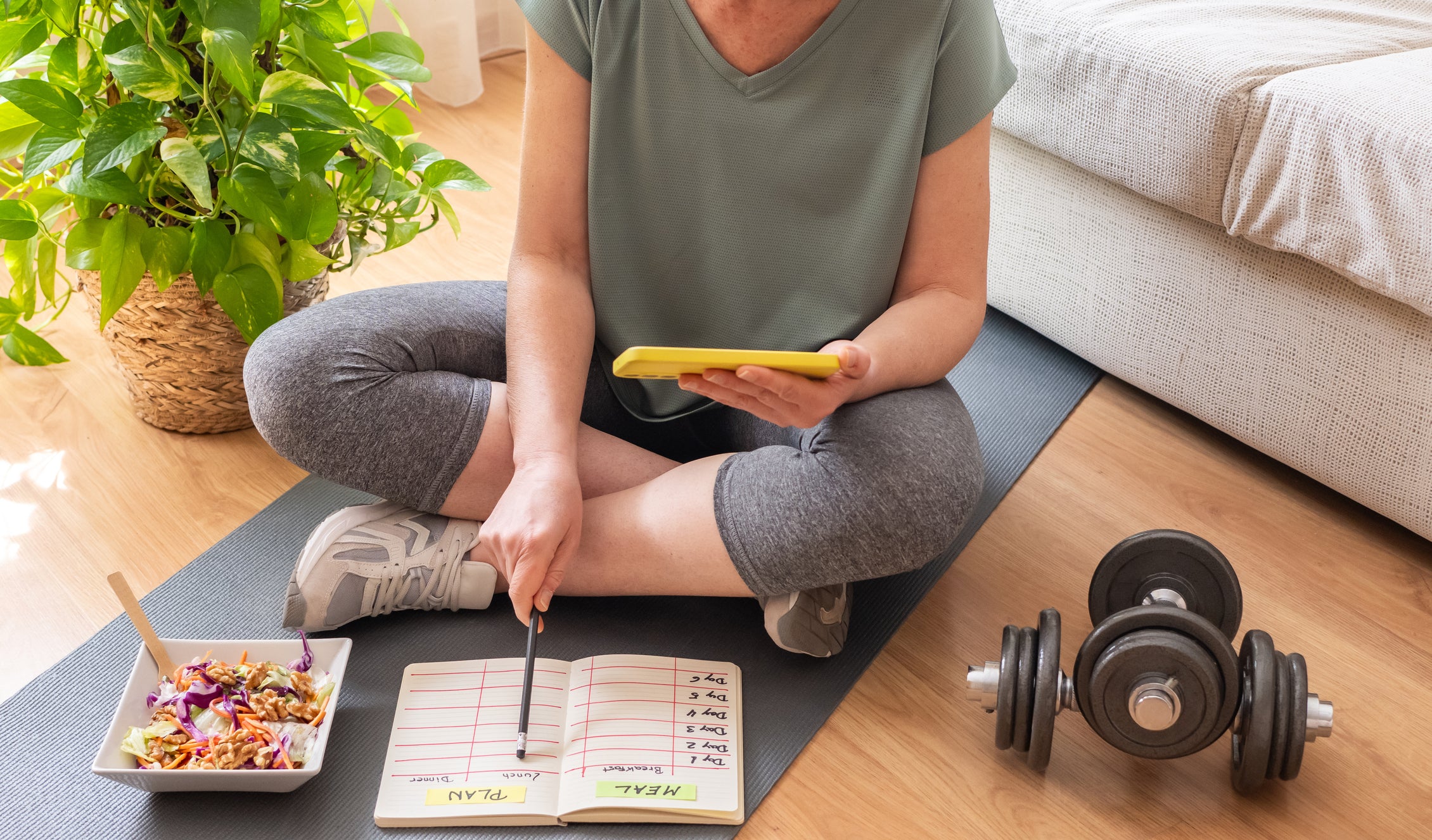 This image shows a person sitting cross-legged on the floor, focused on planning their health and nutrition routine.

They are sitting on a dark exercise mat in a bright, cozy room with wooden floors. A light-colored couch is nearby, and there is a leafy green houseplant to the left, adding a fresh, calm feel to the space.

The person is wearing casual workout clothes—a short-sleeve top, leggings, and athletic shoes. Their face is not visible, but their posture suggests they are relaxed and engaged.

In front of them is an open notebook laid flat on the mat. The notebook is organized into sections labeled “PLAN” on one page and “MEAL” on the other. They are holding a pencil and appear to be writing or tracking something in the notebook.

In their other hand, they are holding a smartphone, likely referencing information such as a meal plan, workout routine, or nutrition details.

To the left of the notebook is a bowl of salad with colorful ingredients like greens and shredded vegetables, suggesting a healthy meal.

To the right, on the floor, there is a dumbbell, representing exercise and strength training.

Overall, the scene conveys a balanced lifestyle—combining nutrition, planning, and physical activity—showing someone actively organizing their daily wellness routine.