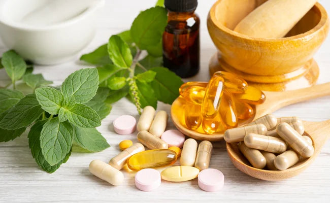 This image shows a collection of herbal and nutritional supplements arranged on a white surface, suggesting natural wellness and traditional remedies.

In the center of the image are several capsules and tablets scattered across the table. The capsules are beige and white, while the tablets are round and pale pink. Nearby are golden softgel capsules, which look smooth and translucent, filled with oil.

To the right is a small wooden bowl filled with capsules, and beside it sits a wooden mortar and pestle, tools traditionally used to grind herbs or medicines. Their presence suggests herbal preparation or natural remedies.

Behind the supplements is a small amber glass bottle, the type commonly used for tinctures or herbal extracts.

On the left side of the image are fresh green mint leaves, adding a natural plant element to the scene. In the background there is also a white bowl, softly blurred, reinforcing the idea of preparing herbs or supplements.